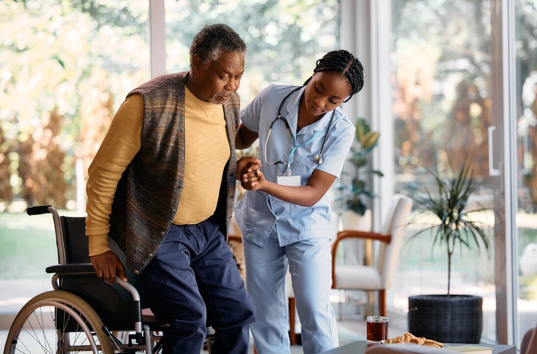 African American senior man getting up from the wheelchair with help of young nurse in nursing home.